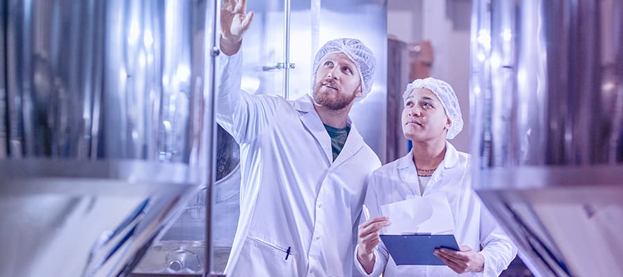 Two life science quality managers wearing lab coats conducting a supplier audit and inspecting equipment at a manufacturing plant.